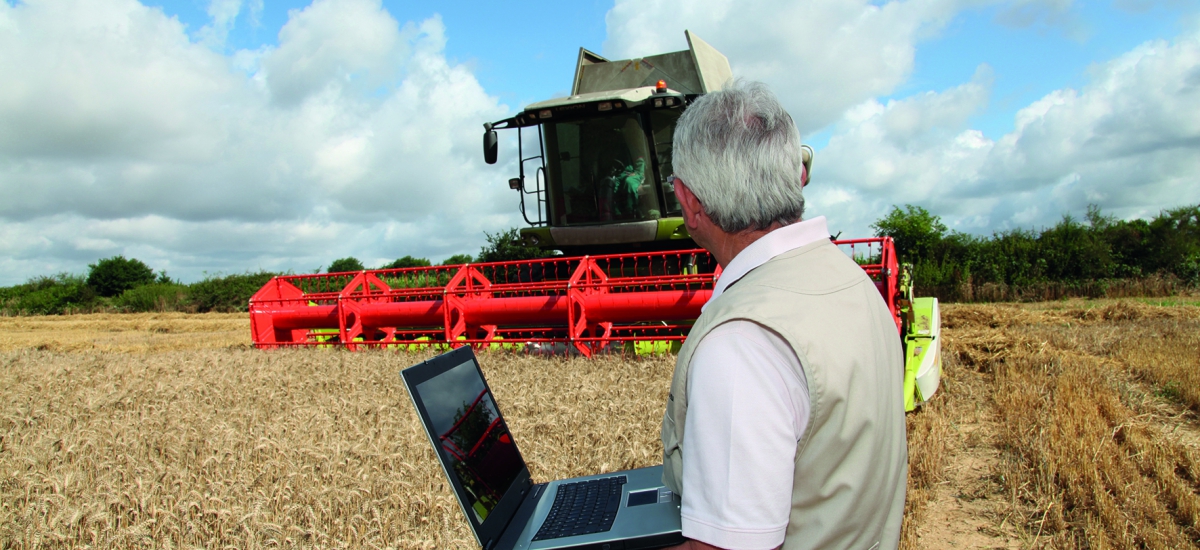 Man in field with laptop, next to a combine.